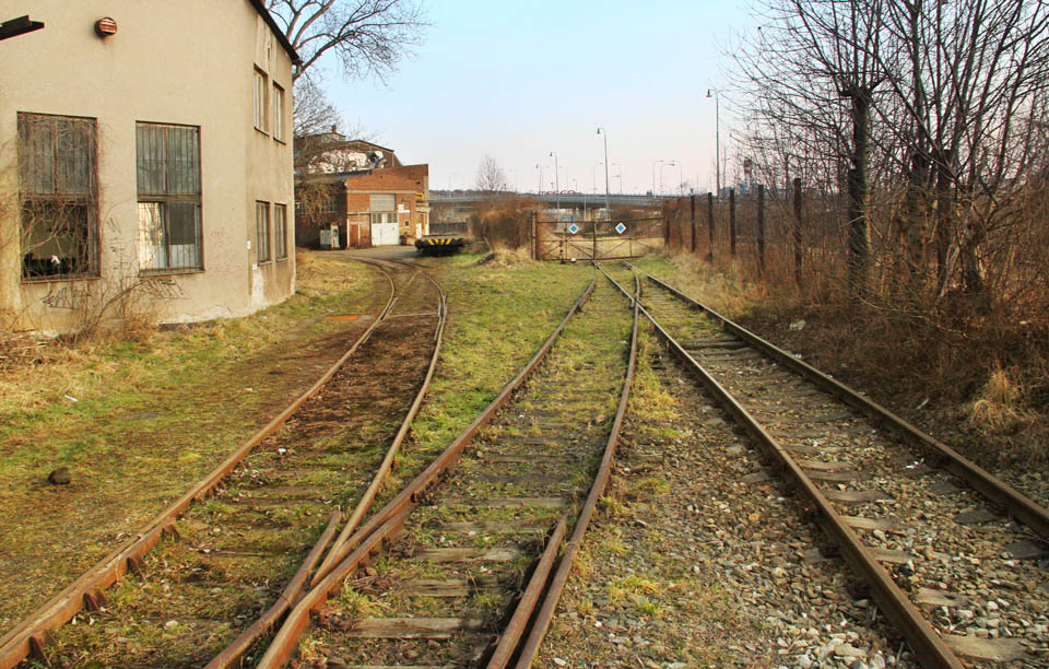 Střídmý pohled za pátou bránu. Dvě koleje vlečky pokračují hlouběji do areálu, jedna se vrací skrz další bránu ven, aby se na chvíli spojila se staniční kolejí před definitivním zvlečkovatěním bývalé dráhy. Střídmý pohled za pátou bránu. Dvě koleje vlečky pokračují hlouběji do areálu, jedna se vrací skrz další bránu ven, aby se na chvíli spojila se staniční kolejí před definitivním zvlečkovatěním bývalé dráhy.