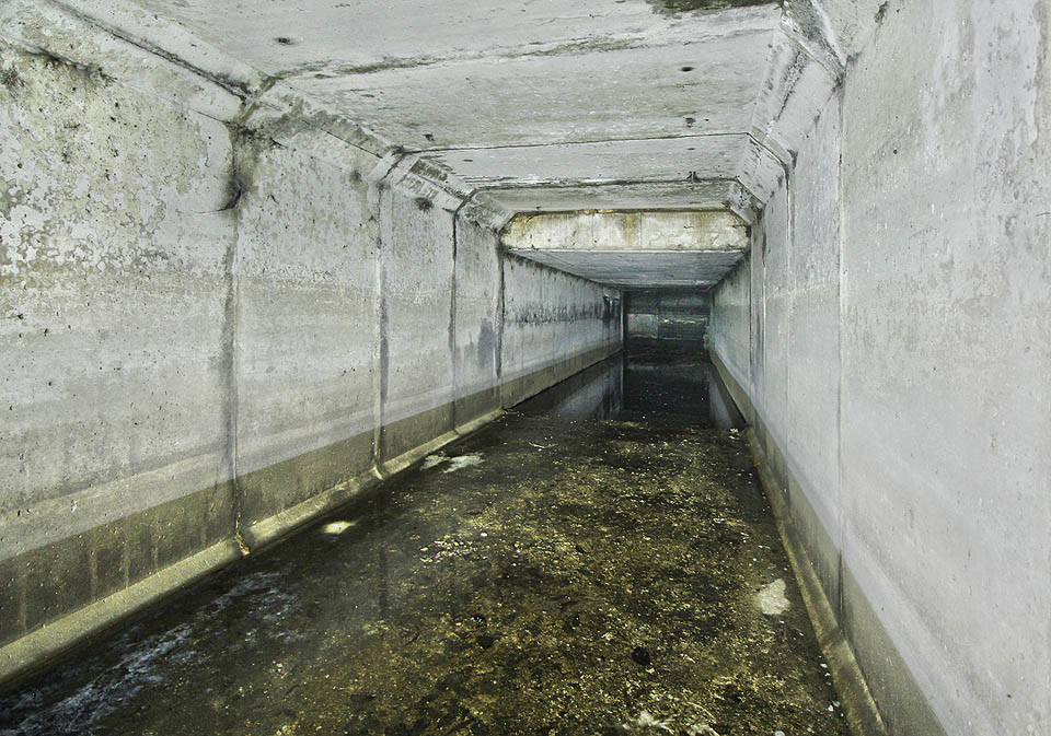 Pokračování spojovací chodby směrem k odlehčovací komoře OKB04. Dobře patrné je snížení stropu pod železniční vlečkou a v dálce zahrazená stěna koryta kmenové stoky B. Pokračování spojovací chodby směrem k odlehčovací komoře OKB04. Dobře patrné je snížení stropu pod železniční vlečkou a v dálce zahrazená stěna koryta kmenové stoky B.
